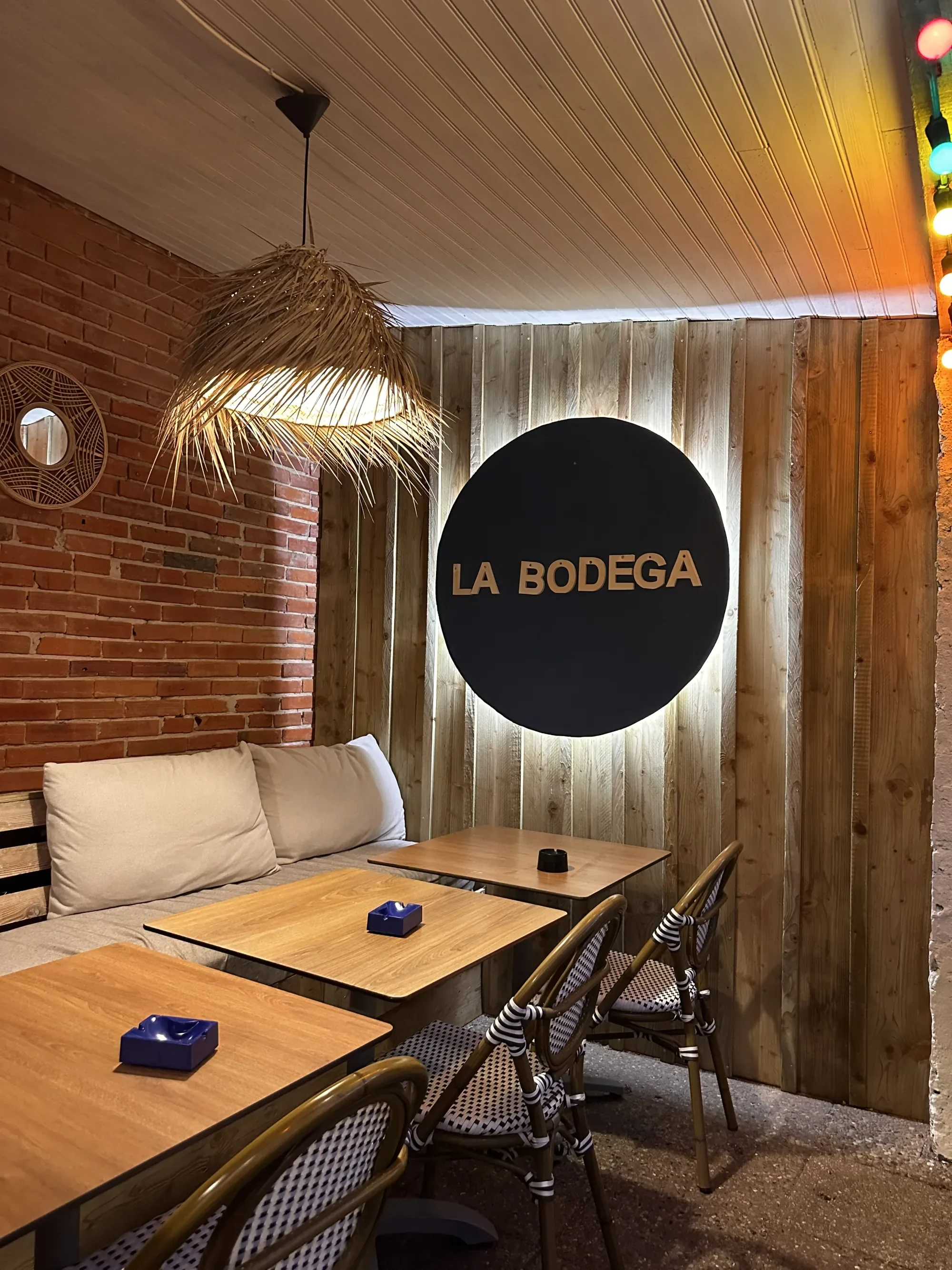 L'intérieur de La Bodega — enseigne ronde noir et or sur lambris bois, banquette et briques